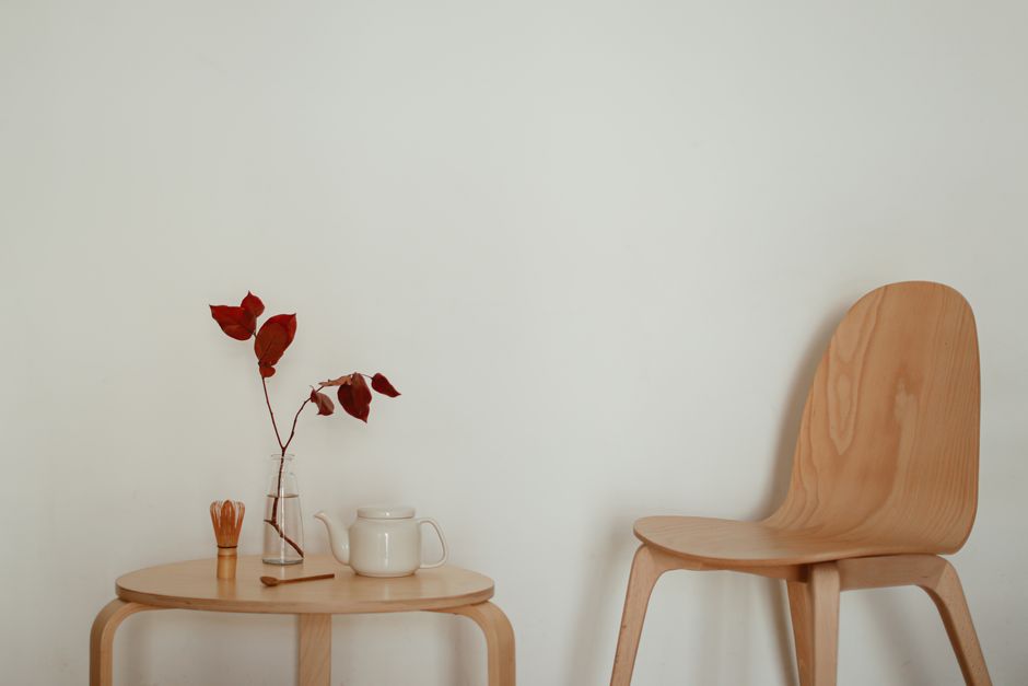 A minimalist interior featuring a table with red leaves in a vase, a teapot, and a wooden chair against a neutral wall.